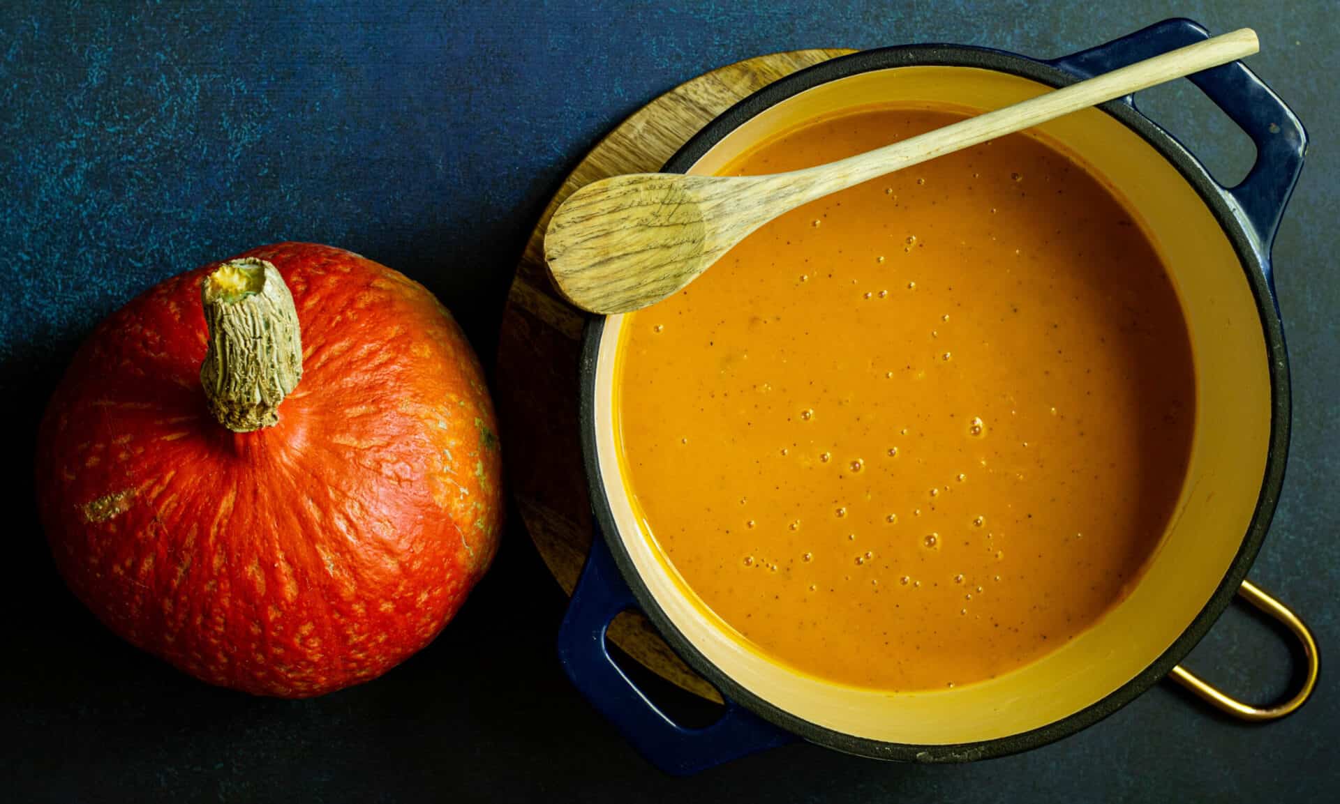 pot of red kuri squash soup on a blue background next to a whole red kuri squash