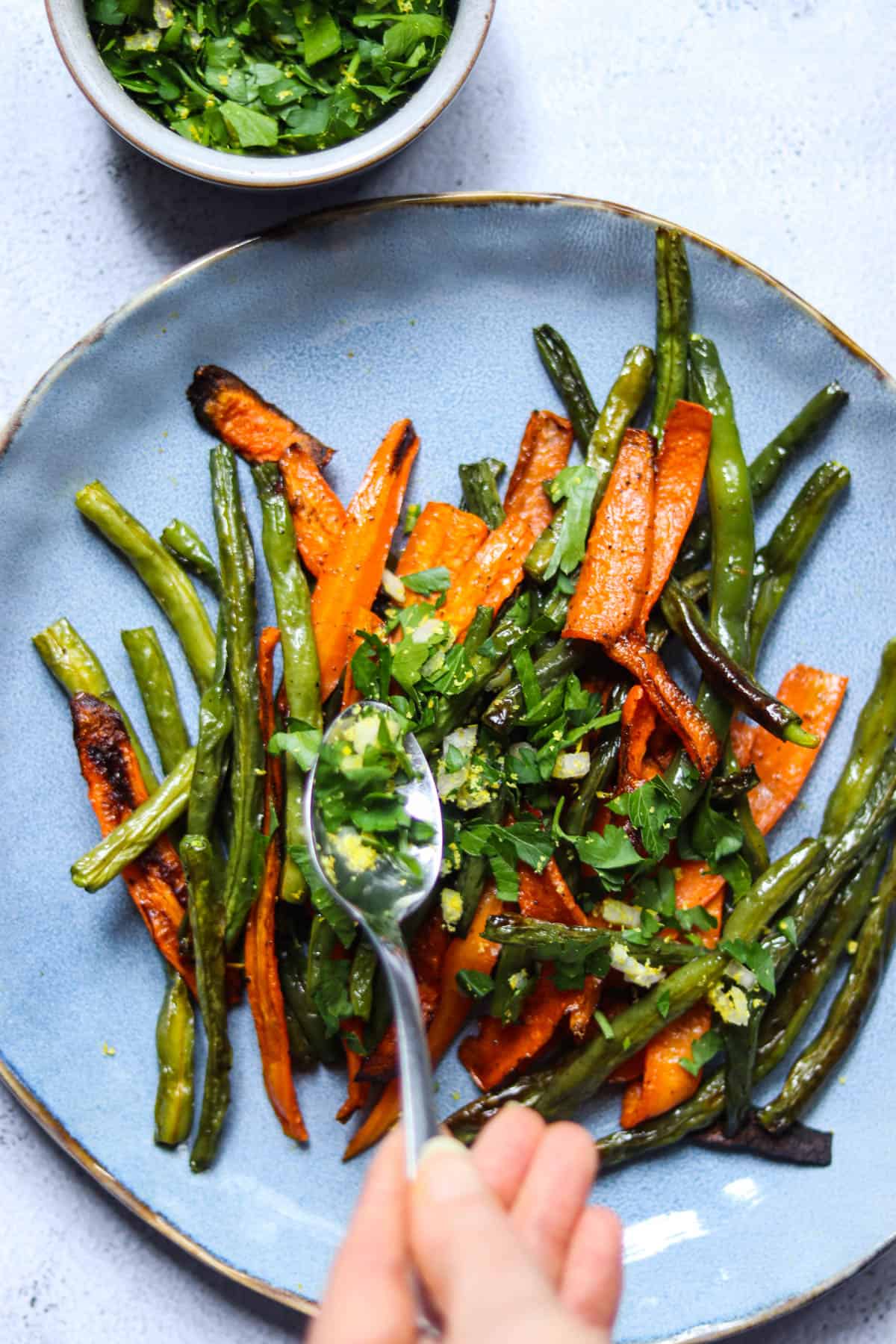 spooning shallot gremolata onto a plate of roasted green beans ad carrots.