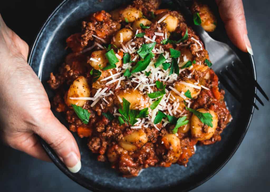 Hands holding a plate of gnocchi and Bolognese sauce.