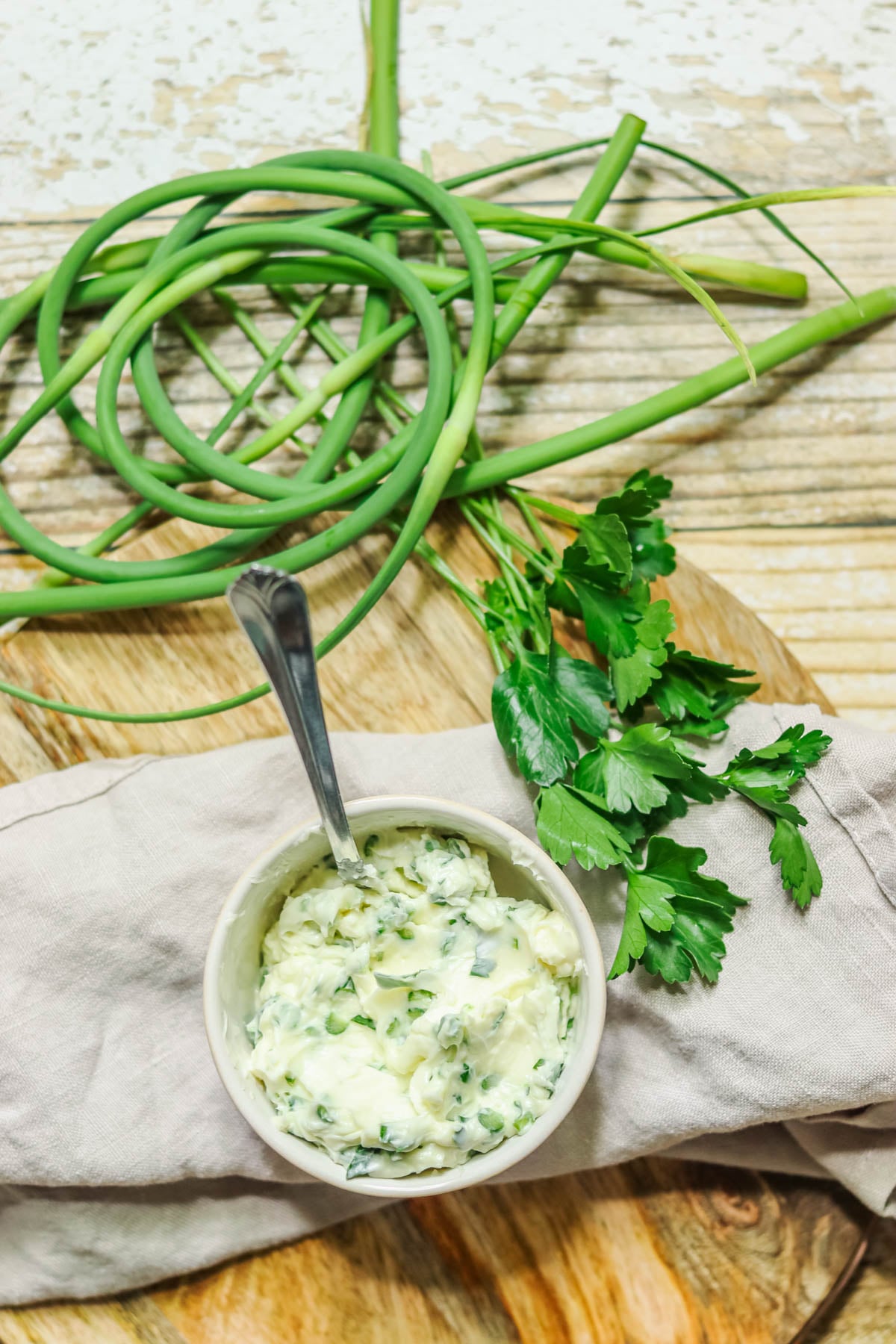 Garlic scape butter in a small bowl sitting on a napkin next to garlic scapes and parsley.